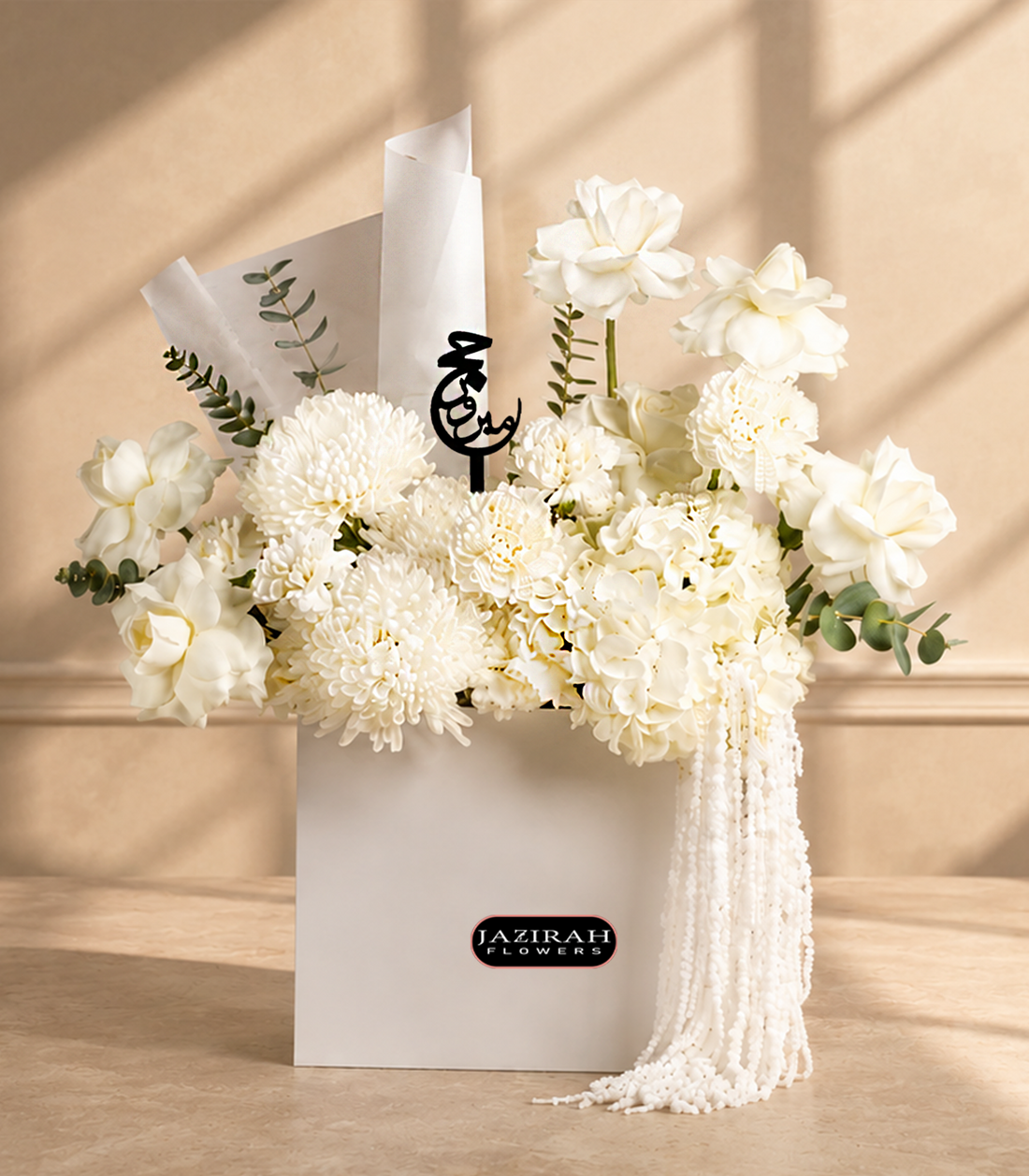 Luxury white flower arrangement for Hajj with roses. hydrangeas, eucalyptus, a black Arabic Hajj Mabroor topper, and pearl fringe in a white gift bag.