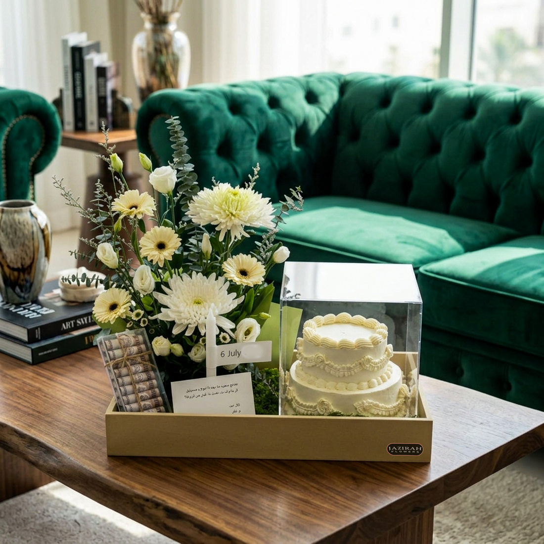 A curated gift set displayed on a wooden coffee table in a cozy living room. The set includes a fresh flower arrangement with white daisies and green foliage, a small white tiered cake under a clear display box, a gift card, and a small metallic item in a tan gift box.