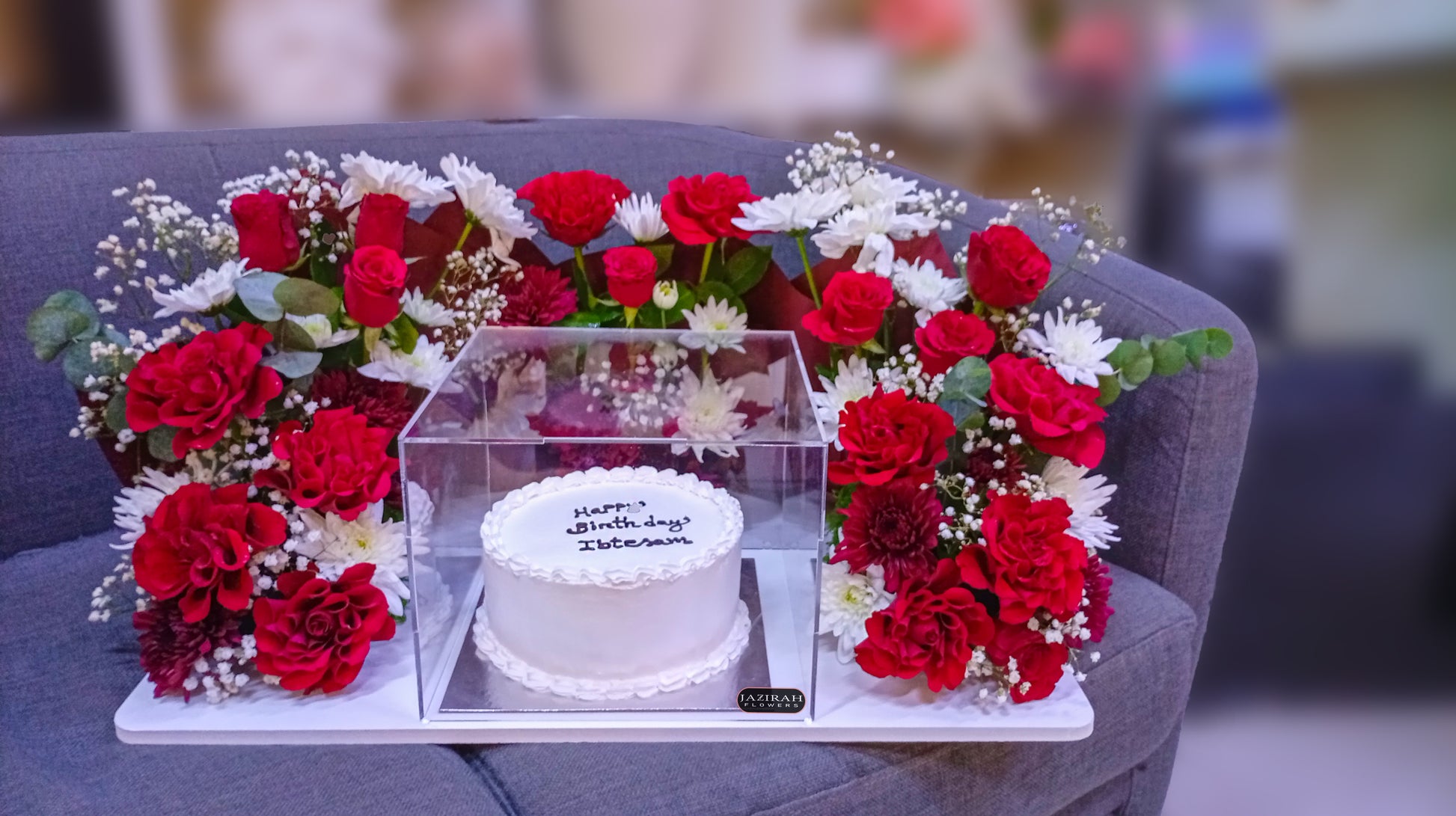 Premium white birthday cake in an acrylic box surrounded by a luxury red rose and white flower arrangement by Jazirah Flowers UAE.