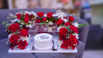 Premium white birthday cake in an acrylic box surrounded by a luxury red rose and white flower arrangement by Jazirah Flowers UAE.
