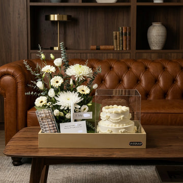 A curated gift set displayed on a wooden coffee table in a cozy living room. The set includes a fresh flower arrangement with white daisies and green foliage, a small white tiered cake under a clear display box, a gift card, and a small metallic item in a tan gift box.