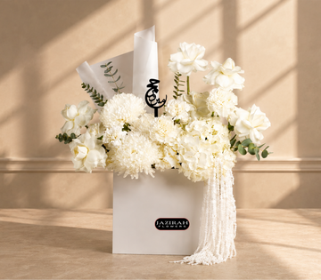 Luxury white flower arrangement for Hajj with roses. hydrangeas, eucalyptus, a black Arabic Hajj Mabroor topper, and pearl fringe in a white gift bag.